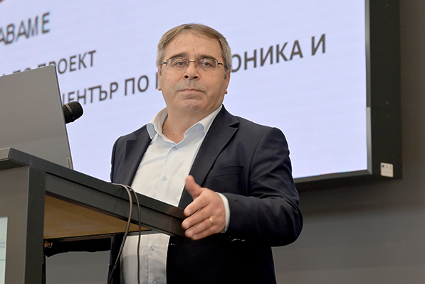 Мъж с очила и тъмно сако говори зад катедра пред голям екран с прожектиран текст
